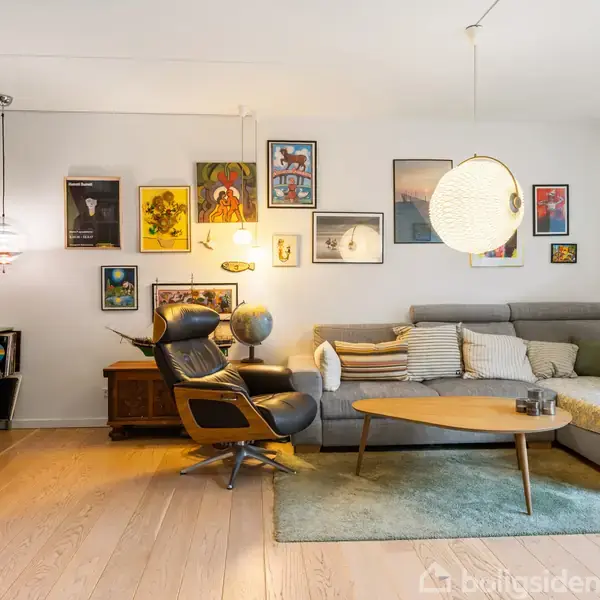 Stue med en grå sofa og et rundt sofabord, omringet af kunst på væggen. En lænestol står ved siden af, nær en lampe og reol. Lyse trægulve og grøn væg.