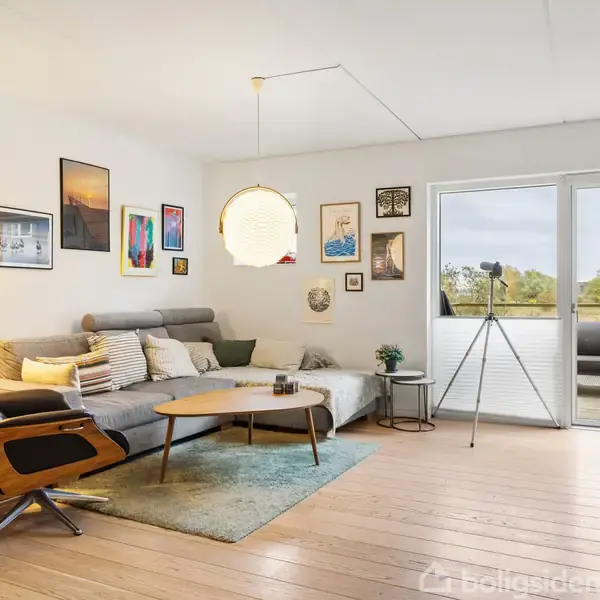 Sofa står i stue med trægulv, flankeret af moderne lænestol og rundt sofabord. Væggene er dekoreret med kunst. En altandør fører til en udendørs terrasse med udsigt over træer.