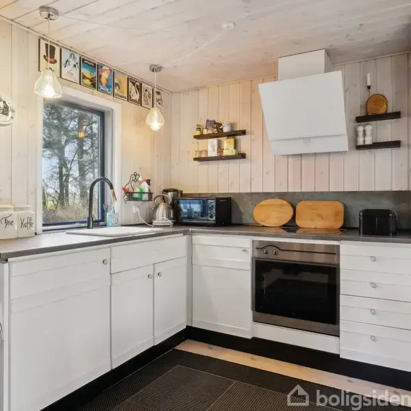 Hvidt køkken med skabe, vask og kogeplader, omkranset af træpanelvægge. En ovn, mikrobølgeovn og diverse køkkenredskaber er synlige. Udendørs udsigt gennem vindue. Tekst på krukker: "Sukker", "Havre", "Kaffe".