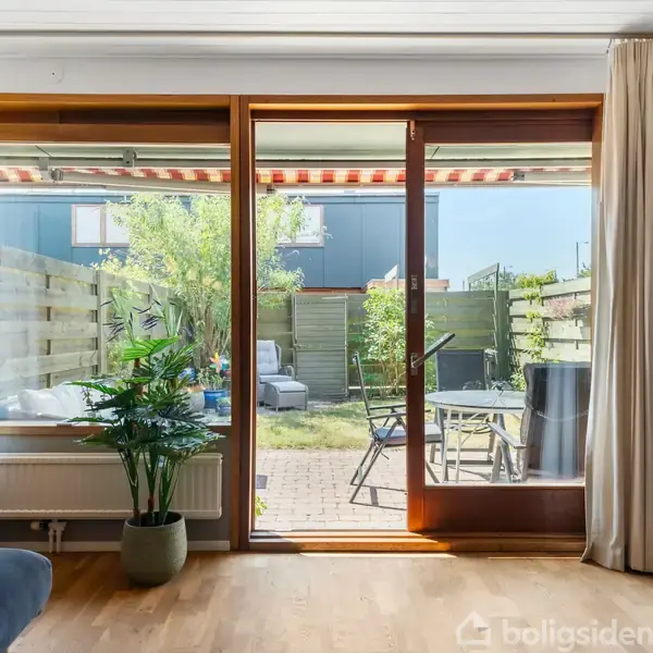 Glasdør åbner mod terrasse med havemøbler og grønne planter. Indendørs ses trægulv, en potteplante og beige gardiner. Udsigt til solrig have med træindhegning.