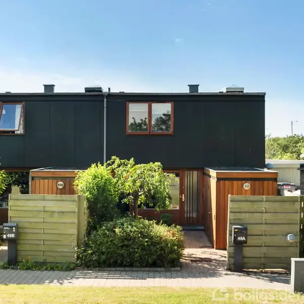 Et rækkehus med sort og træfacade står i en have med grønne buske. To indgange med numrene "46B" og "46A" er synlige på hver side af en fælles sti.