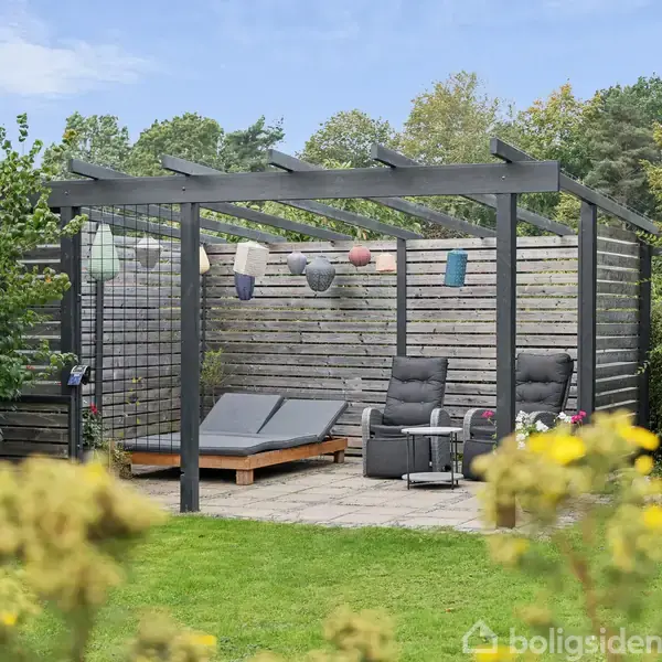 En pergola med hængende lanterner over to hvilestole og en dobbelt solseng. Omgivet af frodig have, med blomster i forgrunden og træer i baggrunden.