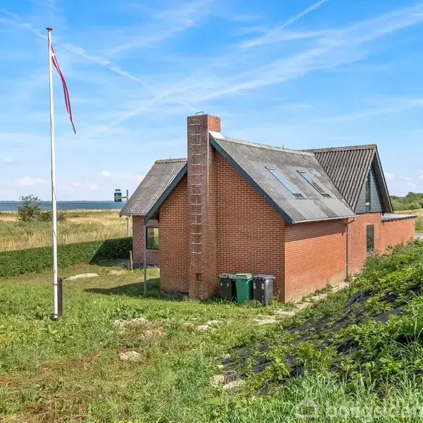Rødt murstenshus med skråtag, står stille. Det er omgivet af en grøn græsplæne og have, nær en flagstang med et flag i stille vind; kysten ses i baggrunden.