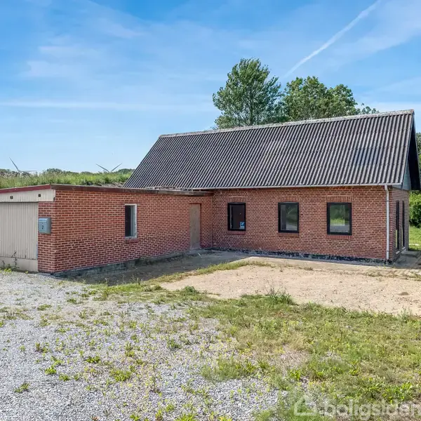 Et rødt murstenshus med sort tag står stille ved siden af en garage, omgivet af en grusbelagt indkørsel og grønne områder med træer i baggrunden under en blå himmel.