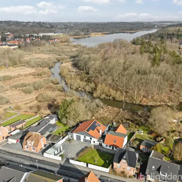 Rækkehuse med røde tage står langs en vej i forgrund, omgivet af træer og en flod, der snor sig gennem et bakket landskab i baggrund.
