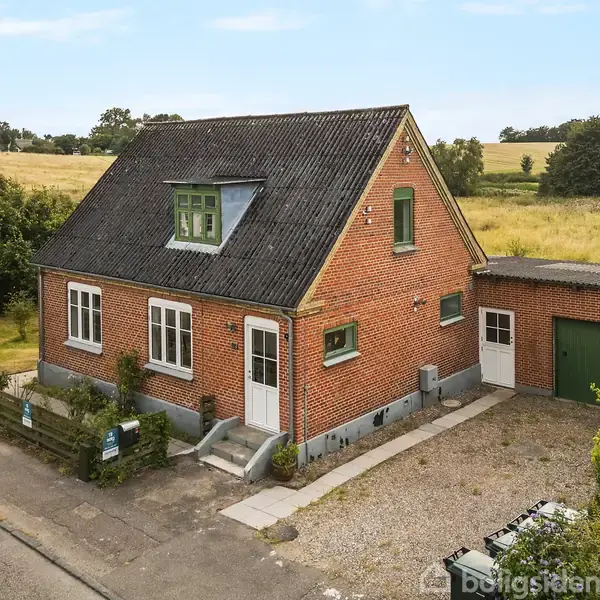 Rødt murstenshus med sort tag og kvist, står ved en vej. Til højre er en garage med grøn port. Baggrunden viser en mark med træer ved horisonten.