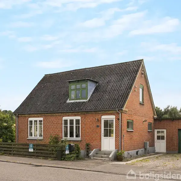 Et rødt murstenshus med sort tag står ved en vej med fortov. Der er en grøn port ved siden af huset og et træ i baggrunden.