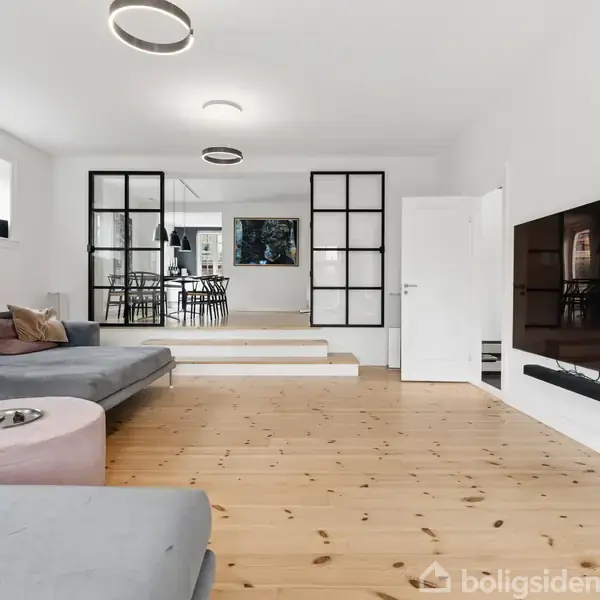 Stue med stor grå sofa og moderne loftslamper, træplankegulve; et fladskærms-tv hænger på væggen. I baggrunden ses en spisestue med sorte stole ved et bord.