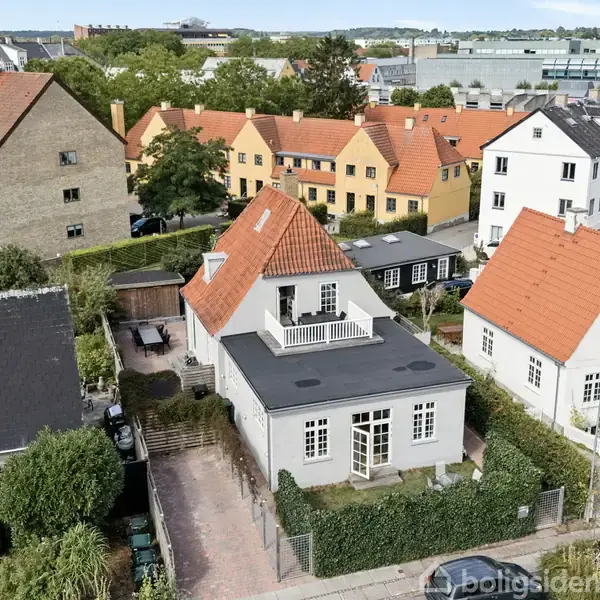 Et hvidt hus med rødt tegltag står i et boligområde omgivet af andre huse. Der er en lille have foran huset og en brolagt sti fører til hoveddøren.