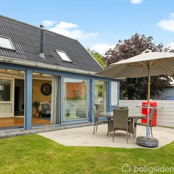 Et hus med store glasdøre åbne mod en terrasse, møbleret med et bord, stole og en parasol i en have. I baggrunden ses en hvid trævæg og træer.