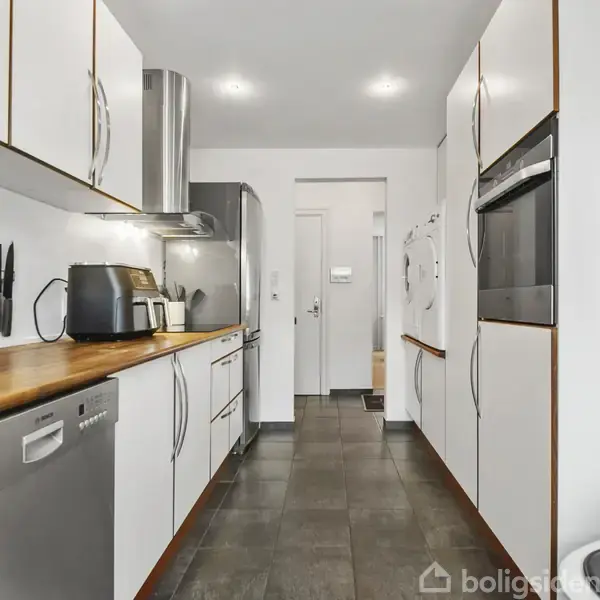 Køkken med hvide skabe og træbordplade, der indeholder en opvaskemaskine, ovn og køkkenmaskiner, såsom en brødrister. Knive er opbevaret i en knivmagnet. Gangen fører til en lys korridor.