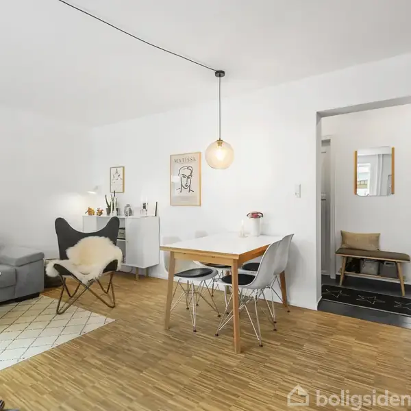 En moderne stue med en grå sofa, et rundt sofabord og en lænestol med pels. En spiseplads med træbord og stole under en pendellampe. Hvidmalede vægge dekoreret med kunstplakater.