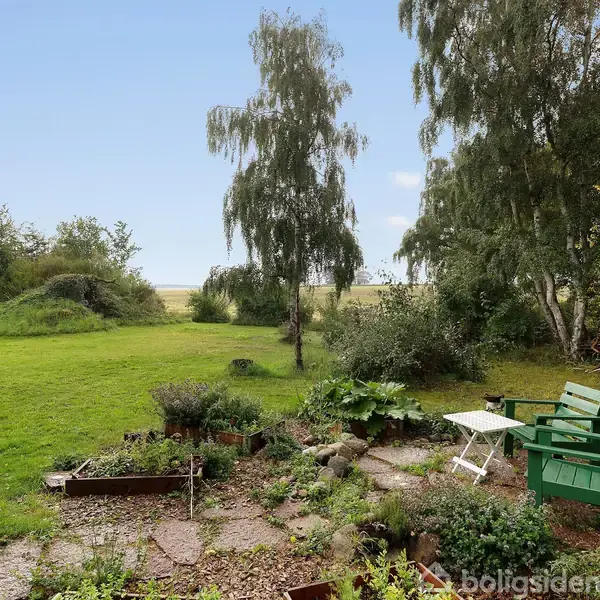 Bænk og bord står stille i en grøn have med græsplæne og træer. Ved siden af er der en brændestabel og blomsterbede.