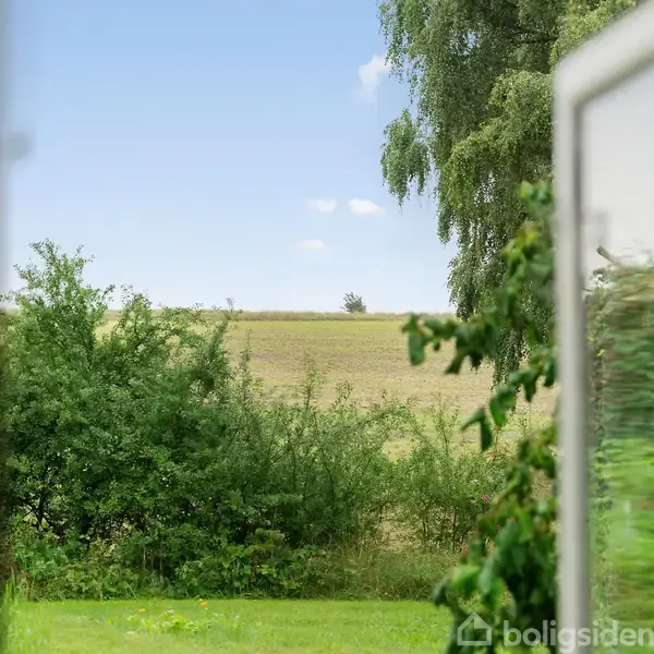 Et åbent vindue viser en udsigt til frodig grøn vegetation og en mark under en klar blå himmel.