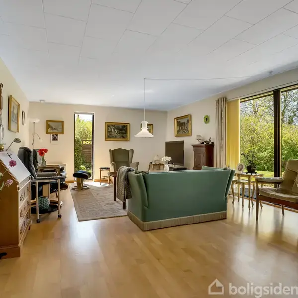 Stor, lys stue med trægulv. En grøn sofa og lænestole omkring et tæppe. Store vinduer med udsigt til have. Der hænger billeder på væggene, og en lampe hænger fra loftet.