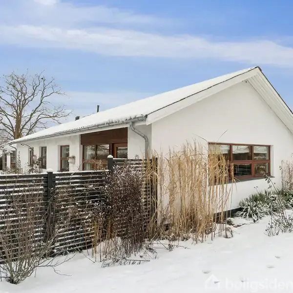 Et hvidt hus står i et snedækket landskab omgivet af en sort træhegn. Buske og træer indrammer ejendommen, og himlen er blå med lette skyer.