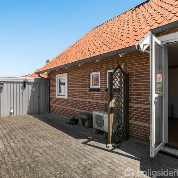 Rød murstensbygning med skråt tegltag. En åben terrassedør fører ud til en flisebelagt gårdhave med en stenterrassepejs til venstre. Blå himmel udenfor.
