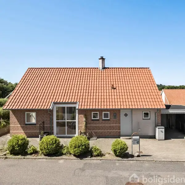 Rødt murstenshus med tegltag står stille på en stille villavej omkranset af hække og buske. Der er en carport til højre og en skorstensrør på taget.
