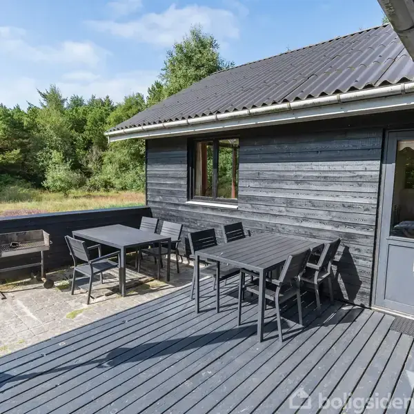 Sortmalet træhus har en terrasse med sort havebord og stole, ved siden af en grill. Omkringliggende grøn vegetation og skov i baggrunden.