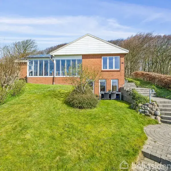 Et rødt murstenshus med store vinduer står på en velplejet, skrånende græsplæne. Til venstre findes en række buske, og til højre en stentrappe omgivet af hæk og træer.