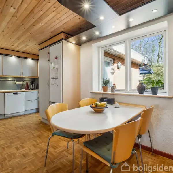 Et ovalt spisebord med gule stole står foran et stort vindue. Køkkenet i baggrunden har hvide skabe og træloft. Parquetgulve fremhæver det lyse rum.