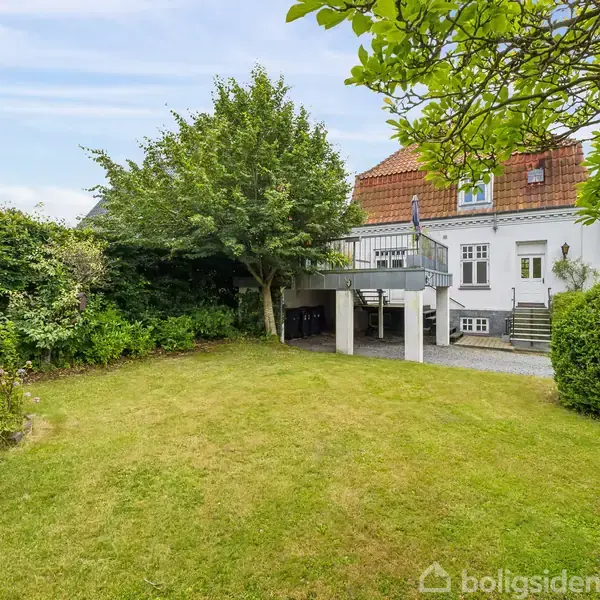Hus med hvid facade og rødt tag; stor terrasse med trappe. Græsplæne omkranset af træer og buske. Drivhus i venstre side.