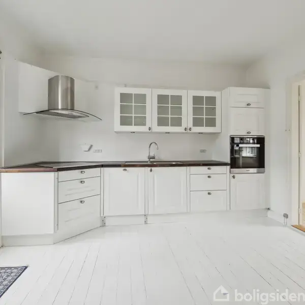 Et hvidt køkken med skabe, ovn, og emhætte står langs væggen. Døren til venstre fører udenfor, og et tilstødende rum ses til højre.