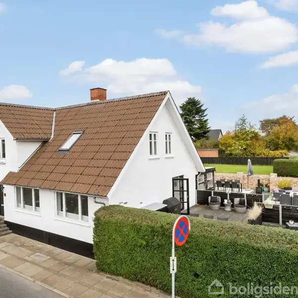 Hvidt hus med brun tegltag, stående på en hjørnegrund omgivet af hække. En åben terrasse med havemøbler ses bagved, mens stien foran er ryddelig. Et skilt markerer parkeringsforbud.
