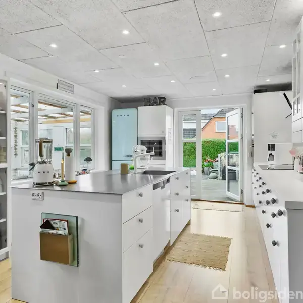 Et lyst køkken indeholder en stor køkkenø med apparater og åbne hylder fyldt med porcelæn. I baggrunden ses en glasdør, der fører ud til en have med buske.