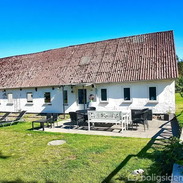 En hvid, langstrakt bygning med rødt tag står i en grøn have. Foran er der en terrasse med havemøbler under en klar blå himmel.
