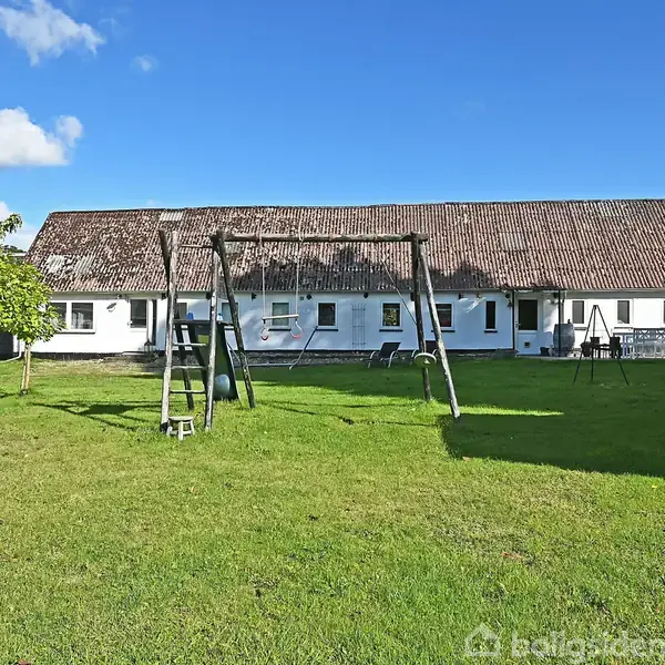 En hvid bygning står i solen med gyngestativ i en grøn have. Et lille træ vokser ved siden af under en blå himmel med skyer.