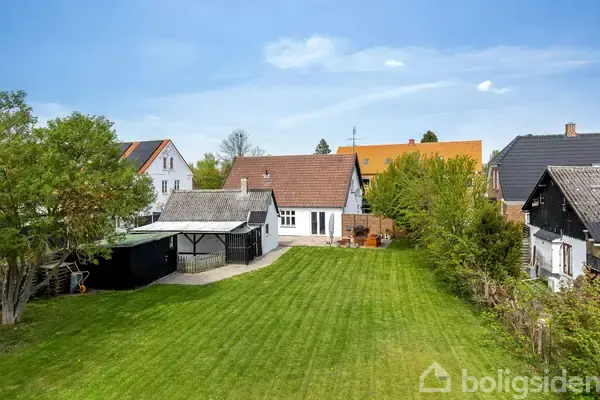 Hvidt hus med brunt tag står i en have med velplejet græsplæne. Ved siden af huset er der et skur og en indkørsel, omgivet af andre huse og grønt løv.