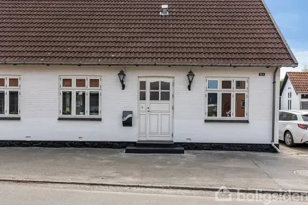 En hvid murstenshus med en rødbrun tegltag, symmetrisk placerede vinduer og en hvid hoveddør med glaspaneler. Huset står på en stille gade med parkerede biler.