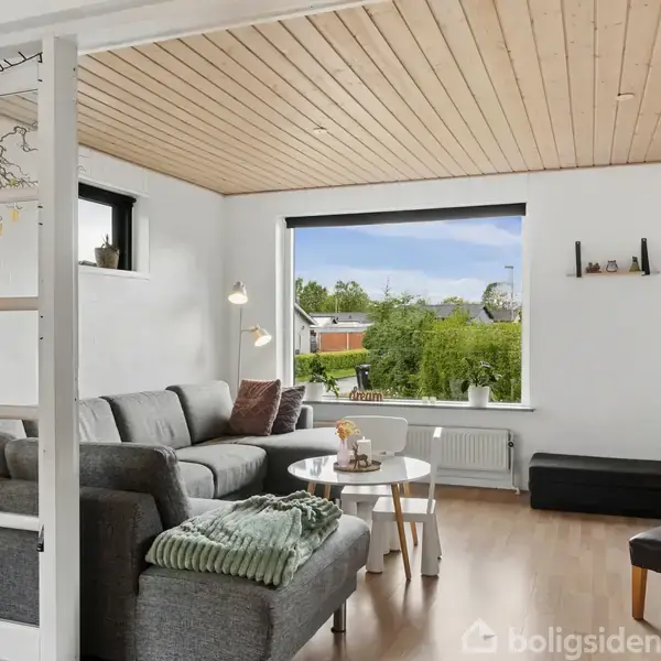Stue med grå hjørnesofa, sofabord med blomster, og puder, set mod vindue med udsigt til grøn have. Loft og gulv er i lyst træ, hvide vægge, og hylder til pynt.