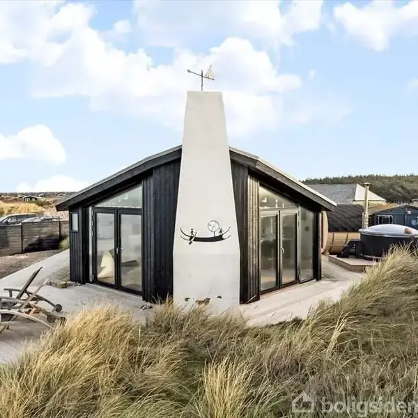 En sort træbygning står stille omgivet af sand og græs, med en brændeovn i midten. Den har store glasvinduer, og borde samt stole står på terrassen.