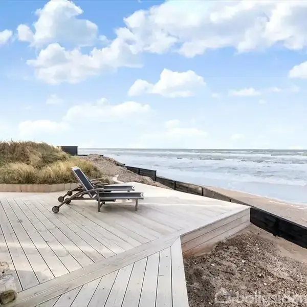 Terrasse med to liggestole og havudsigt. En pejs til venstre tilføjer hygge. Omgivet af strand og bølger under en blå himmel med skyer.