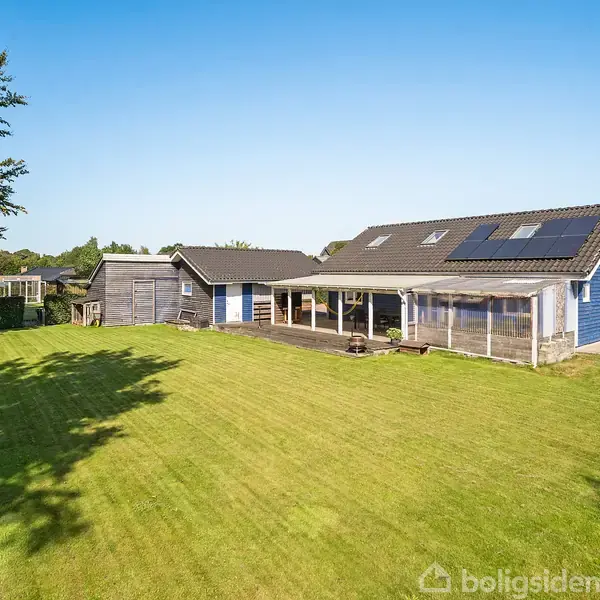 En moderne, blå etplansvilla med solpaneler på taget står i en velplejet have med stor græsplæne og omgivende træer under en klar blå himmel.