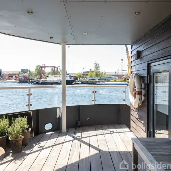 Udsigtsplatform med glasrækværk, livredningskrans og nogle planter i krukker; ombord på en båd med udsigt til en havn med bygninger og kraner i baggrunden.