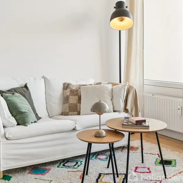 Sofa med puder står stille ved et vindue. To runde træborde har en lampe og bøger på sig. Lysestander oplyser krog i moderne stue med farverigt tæppe.