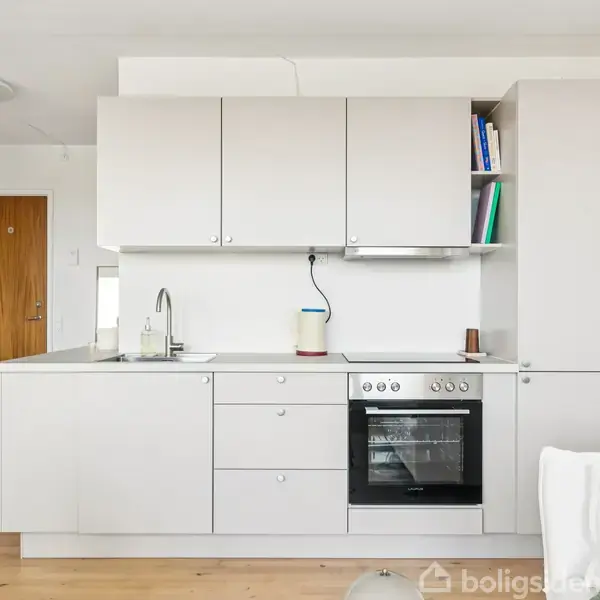 Et moderne køkken med lysegrå skabe og skuffer, en indbygget ovn og et blankt køkkenbord. Til venstre ses en trædør og en tæppebelagt gang i en lys lejlighed.