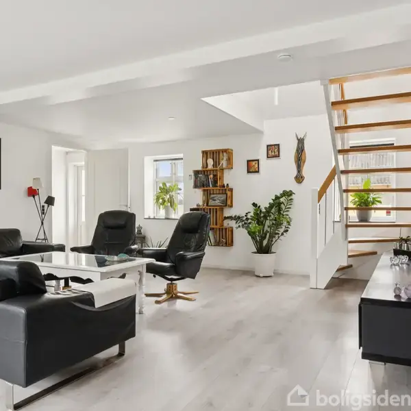 Sort sofaarrangement i moderne, lyst stueområde. Lænestol ved glasbord, trappe til højre. Planter og dekorationer pryder væggen. To vinduer giver naturligt lys. Tekst på væggen: "Home Sweet Home".