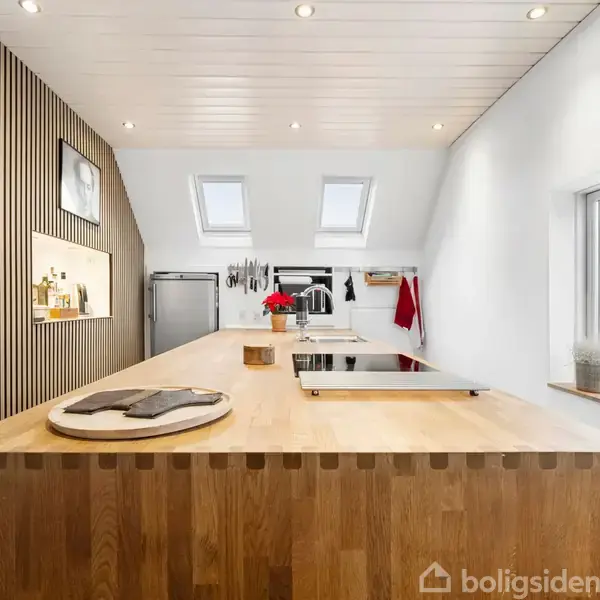 Et stort køkkenbord i træ dominerer rummet, med indbyggede kogeplader på toppen. Kontekst: Moderne køkken med ovenlysvinduer, planter på vindueskarmen og hylder på væggen.