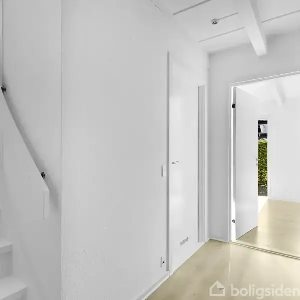 En hvid trappe fører opad ved siden af en gang med lyse trægulve. Døren for enden af gangen åbner ind til et rum med en udsigt til grøn vegetation.