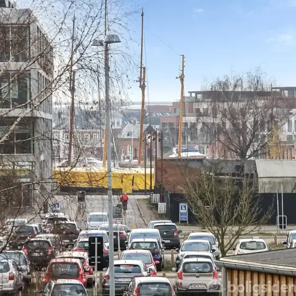 En række parkerede biler står stille på en byvej omgivet af moderne bygninger og træer. Personer går på fortovet ved siden af vejen, og der ses master i baggrunden.