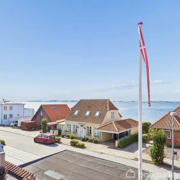 Flagstang med rød-hvidt flag vajer i forhaven til en række huse, der ligger ved en vej nær kysten. Blå himmel og et kig mod havet i baggrunden.