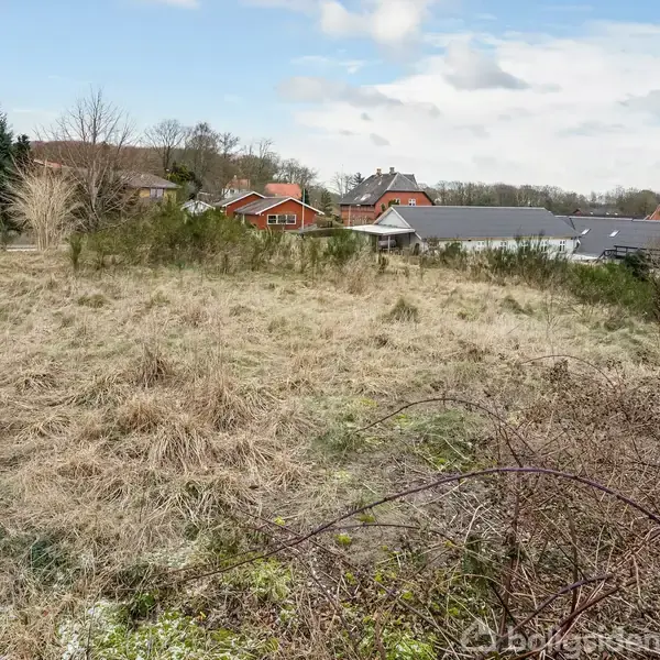 En ubebygget grund med tørt, vildt græs. I baggrunden ses huse med røde tage og et gennemgående boligområde. Skyer og blå himmel danner himmelbaggrund.