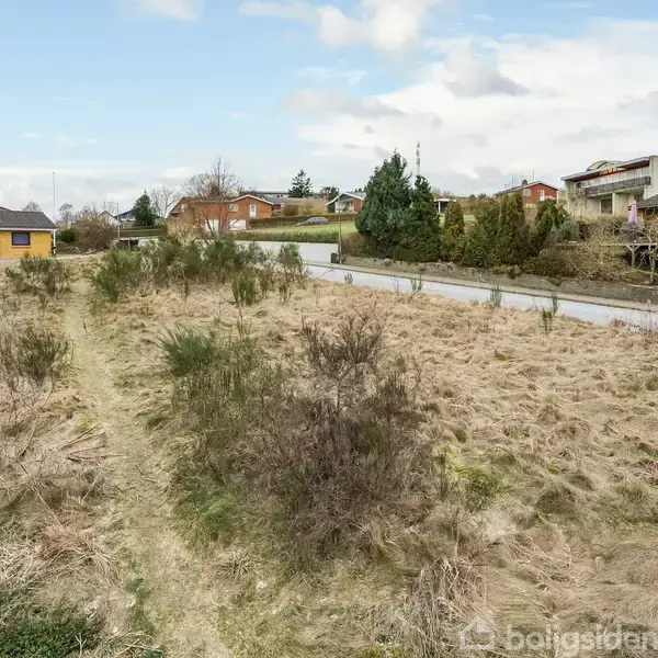 En mark med tørt græs og buske strækker sig ud, med en gangsti i midten. Omkringliggende huse ses i baggrunden på en solrig dag med få skyer på himlen.