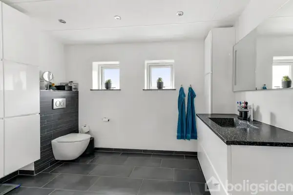 Badeværelse med sort gulv og hvide vægge. Hængende toilet mod venstre med grå fliser. To håndklæder hænger under to små vinduer. Til højre en vask med spejl.