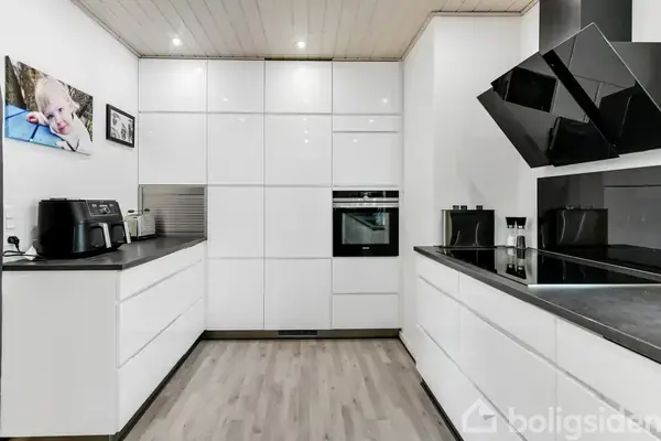 Hvid moderne køkkenindretning med indbyggede skabe og apparater. En ovn er integreret i skabsvæggen. En airfryer står på bordet. Trægulv og hvid loft med indbyggede spotlys. Billede af et barn på væggen.