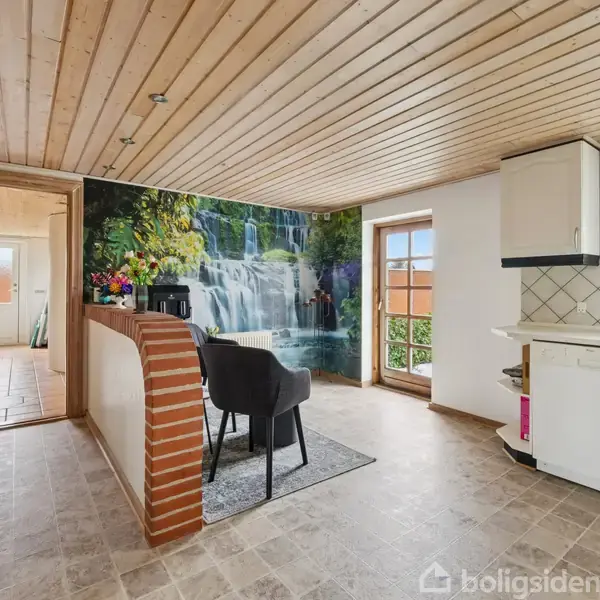 Et køkken med en fototapet af et vandfald på væggen, to sorte stole ved et lille bord, træloft og flisegulv; dagslys skinner ind gennem store vinduer.